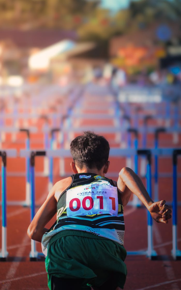 A young runner with numbered bib preparing for a hurdle race on a sunny track.